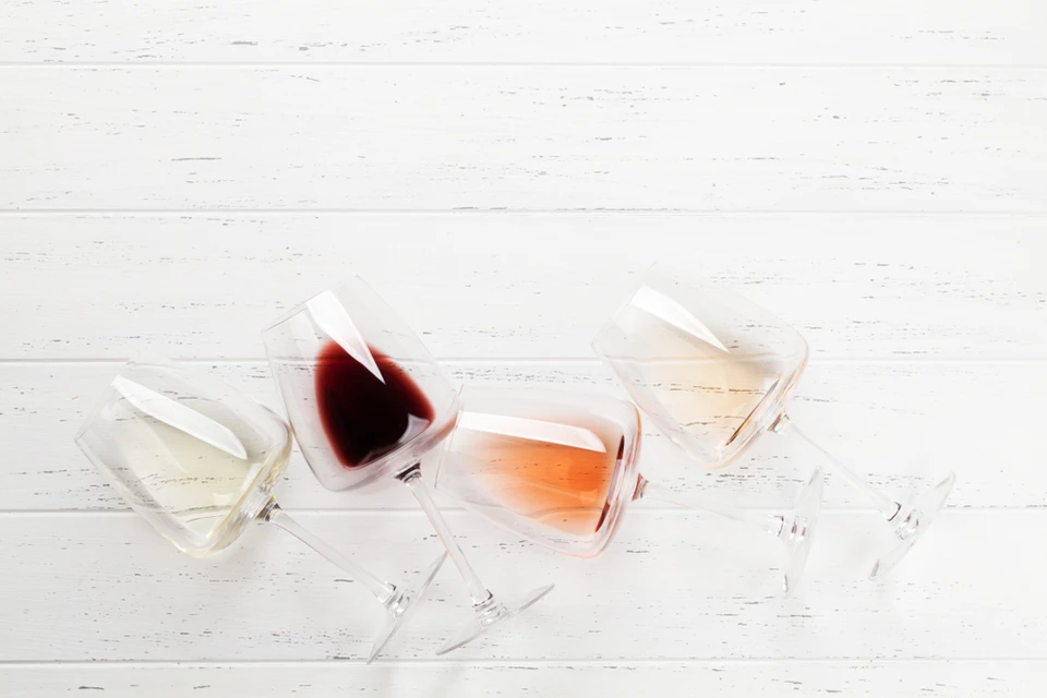 Quatre verres à vin renversés sur surface en bois blanc patiné, contenant différents types de vins : blanc, rouge foncé, rosé et champagne