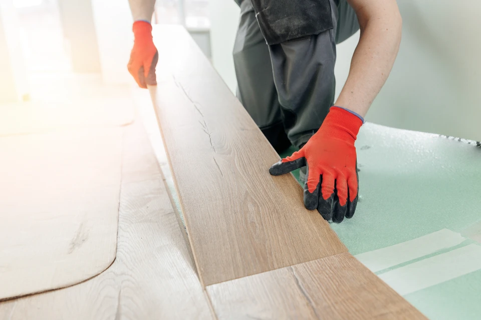 Homme installant du parquet en bois dans une pièce blanche, agenouillé sur le sol, positionnant des lames de parquet avec ses mains
